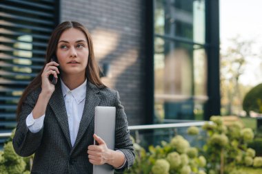 Genç, meşgul bir iş kadını elinde dizüstü bilgisayarla şehir caddesinde yürüyor, telefonla konuşuyor. Başarılı bir iş adamı