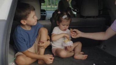 Aç çocuk ve kız aile arabasının bagajında lezzetli atıştırmalıklar yiyorlar. Araba yolculuğu kavramı