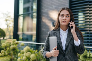 Kendine güvenen iş kadını dizüstü bilgisayarını tutuyor ve modern binanın arka planında telefonla konuşuyor. Uzayı kopyala Metin için boş alan.