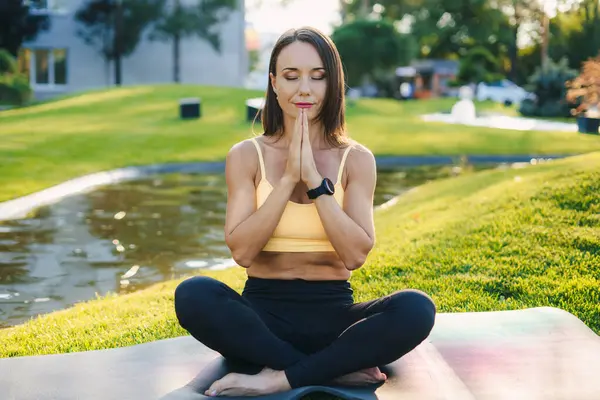 Beautiful adult woman doing breathing yoga exercise in the summer park. Caucasian female meditation pose, healthcare.