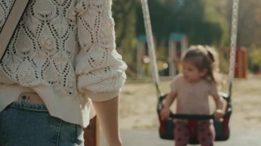 Mutlu küçük kız salıncakta salıncak sürüyor ve parkta etrafı seyrediyor. Sevimli çocuk, kızını sallayan ve anaokulunda ya da boş zamanlarında eğlenen kadın.