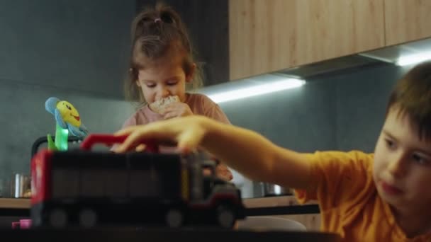 姉のそばに座っているキッズボーイは 屋内のキッチンでテーブルの上のおもちゃトラックで遊んでいました ハッピーファミリー キッズドリーム教育コンセプト — ストック動画