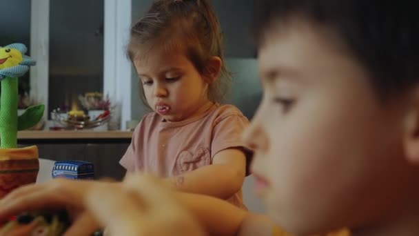 キッチンテーブルで兄と遊んでいるときに おかしな小さな赤ん坊の女の子が口でいっぱいを食べたり噛んだりする 愛らしい幼児の幼児は食べ物をチェックする — ストック動画