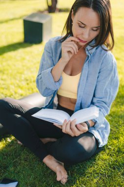 Güneşli bir günde şehir parkında yeşil çimlerde oturan ilginç kitap okuyan çekici yetişkin kadın. Boş zaman kavramının tadını çıkarıyorum..