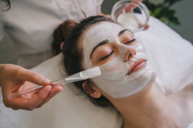 Profesyonel kozmetik uzmanı bir kadının yüzüne nemlendirici maske takıyor. Çekici genç bayan cilt bakımı yaptırıyor..