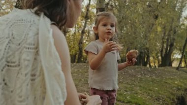 Küçük kız, annesiyle birlikte gölün kenarında durmuş yaz parkındaki ördekleri beslemek için hazırlanmış bir dilim ekmekten yiyor. Mutlu çocuk gözlemleyerek eğleniyor.