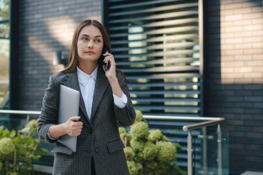 Kendine güvenen bir girişimci müşterisiyle telefonda konuşuyor. Sokakta dikilirken dizüstü bilgisayarını tutuyor. Genç ve meşgul bir iş kadını. Otantik çekim