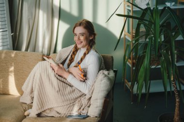 Kızıl saçlı kadın kameraya bakıp gülümsüyor, evde kanepede otururken notlar yazıyor ve düşünüyor. Odaklanmış, tatlı bir kadın...