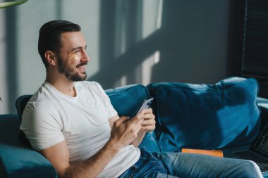 Evde kanepede otururken akıllı telefon kullanan yakışıklı, rahat bir adam. Arkadaşlarla mesajlaşıyorum. Keyifli hafta sonu.