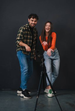 Sıradan giyinen, çağdaş dans eden, dans figürlerini gösteren, cep telefonunu tripodun üzerinde kullanan, siyah arka planda duran genç bir çift. Beyaz ırk