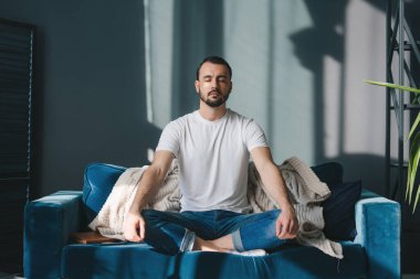 Beyaz tişörtlü beyaz bir adam, lotus pozisyonunda oturuyor, gözleri kapalı meditasyon yapıyor, evdeki kanepede oturuyor. Denge kavramı, uyum, rahatlama.