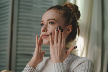 Genç kızıl kadın yüzüne dokunuyor. Gözlerinin altından şakaklarına kadar parmaklarını hareket ettiriyor. Güzellik portresi ve cilt bakım ürünleri ticari çekim