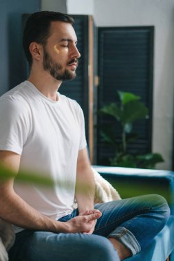 Günlük giysiler giyen, yoga egzersizleri yapan, evdeki kanepede oturan, oturma odasında boş zaman geçiren genç bir adam. İç dengeyi bulmak, stresi yönetmek.