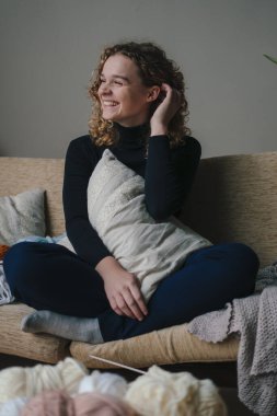 Gündelik kıyafetlerle gülümseyen genç bir kadın sarılan bir yastık giyer, keyifli bir ifadeyle kanepede oturur, oturma odasında dinlenir, başka tarafa bakar. Modern mobilya