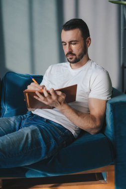 Millennial 'daki adam oturma odasında kanepede otururken not alıyor ya da liste yapıyor. Beyaz adam..