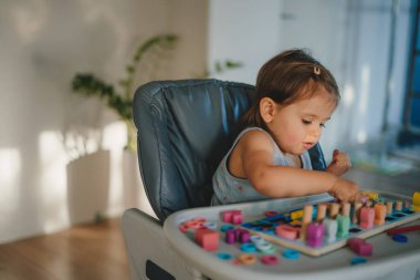 Tahta şekilli, rakamlarla, harflerle oynayan küçük bir kız oyun odasındaki masada oyuncak ayırıyor. Bebekler için eğitim kurulları. Oynayarak öğrenmek.