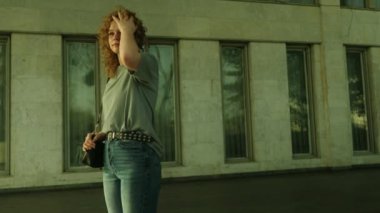 Cana yakın, genç kıvırcık saçlı, t-shir giyen, Jenna, dışarıda, pencereleri olan bir bina duvarında beklerken rahatlıyor. Boş alan