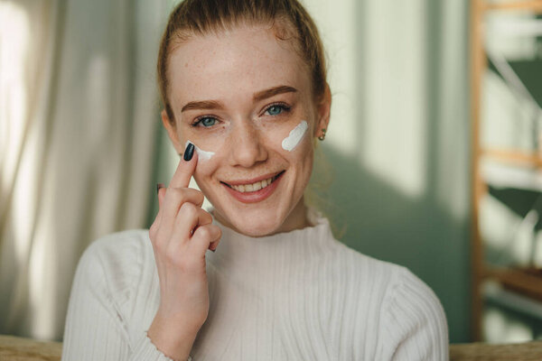 Портрет молодой кавказки в уютной одежде, сидящей на диване и смотрящей в камеру, нанося крем на лицо. Рыжая девушка с волосами заботится о коже. Концепция