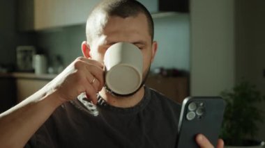 Mutfakta kahvaltı yapan, çay içen ve telefonla mesaj gönderen bir adamın ön görüntüsü. Sosyal ağ, mesaj, teknoloji kavramı.