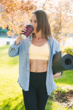 Esmer saçlı, güzel, beyaz bir kadın parkta yoga minderiyle durup bir fincan çay ya da kahve içiyor..