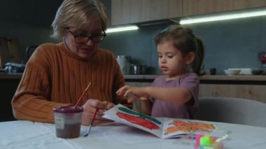Küçük kız evde suluboya ve guaj ile resim yapıyor, büyükannesiyle birlikte, masada oturuyor. Çocukların yaratıcılık kavramı.