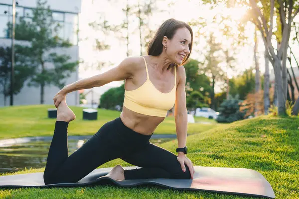 成熟したプロの女性は 夏の朝の公園でヨガマットに座って柔軟な運動をしている彼女の体を伸ばします 女性のフィット感と健康 ロイヤリティフリーのストック画像
