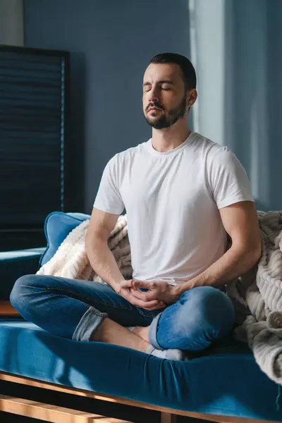 瞑想を通じて健康的なライフスタイルを促進する柔らかいソファの上に座った居心地の良い服装の白人男性 ハーモニーと禅 ストック画像