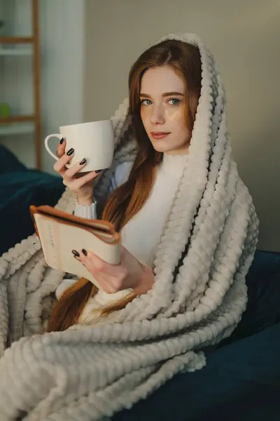 朝早くから美しいジンジャーの若い女性 本を読んで 快適なソファーに座ってカップからホットコーヒーを楽しんでいます 居心地の良い国内の雰囲気 ストック写真