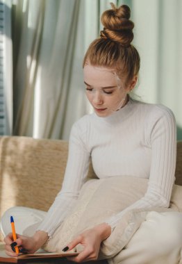 Yüzünde kozmetik maske örtüsüyle evdeki rahat sandalyeye oturmuş kağıt defterde notlar alan neşeli genç bir kadın. İçerideki yaşam tarzı