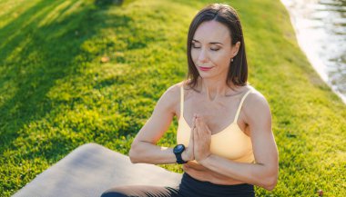 Yetişkin kadın eğitmen, şehir parkının dışında yoga minderinde nilüfer pozisyonunda oturuyor. Namaste Mudra, oturmuş yoga asana, nilüfer çiçeği pozu, nilüfer çiçeği duruşu, Padmasana, çapraz