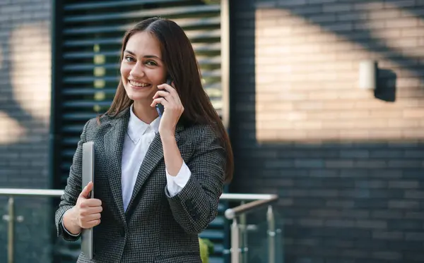 Happy young professional caucasian woman with laptop making a call against office building backdrop. Young cheerful stylish businesswoman. Freelance concept