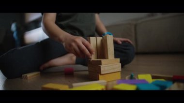 Çocuk ahşap zeminde oturur, tahta bloklarla oynar. Baş parmakları ahşap lekelere bastırarak sanat yaratıyor. Rahatlık ve dinlenme odayı doldurur.