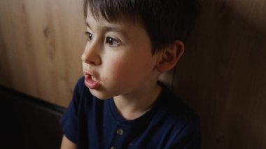 Küçük bir çocuk tahta bir duvarın yanında oturuyor, dilini çıkarırken burnunu ve yanaklarını şakaya vuruyor. Bulaşıcı gülüşü odayı aydınlatıyor.