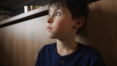 Genç bir çocuk tahta bir duvara yaslanıyor, yukarı bakıyor. Çocuk etrafını keşfederken sahne bir merak ve masumiyet hissi yayıyor.