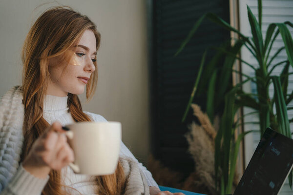 Young woman working on laptop while drinking a cup of tea sitting on the sofa at home. Work from home at cozy atmosphere concept