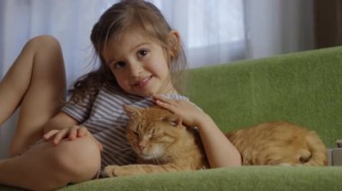 Genç bir kız yeşil bir kanepede tüylü turuncu kedisine sarılırken neşe ve sevgiyle ışıldıyor.