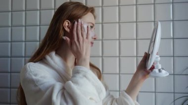 Rahat bir bornoz giyen bir kadın aynanın önünde dikilirken en sevdiği cilt bakım ürünlerini uyguluyor.