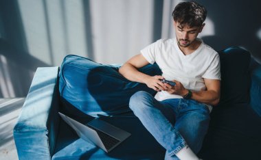 Genç bir adam mavi bir kanepede rahat bir şekilde oturuyor, parlak, güneşli bir odada akıllı telefonuyla derinden meşgul.