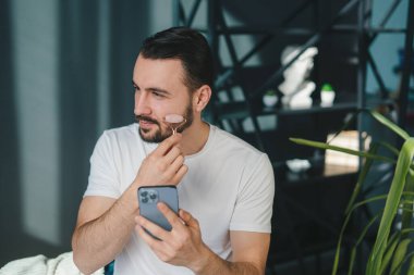 Bir adam akıllı telefonunu kontrol ederken bir cilt ürününü uygular, modern erkeklerin bakım rutinlerini gösterir..