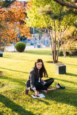 Genç bir kadın neşeli bir şekilde açık havada canlı sonbahar ağaçlarıyla çevrili yumuşak çimenlere çizimler yapıyor.