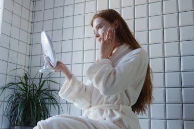 Bir kadın şık bir banyoda rahatlarken aynada tenini incelikle inceler.