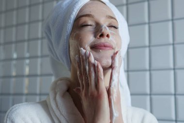 Bir kadın ferahlatıcı bir cilt bakım rutininden zevk alır, parlak bir banyoda yüzünü köpükle temizler.
