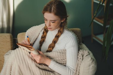 Bir kadın battaniyeye sarılmış, defterine bir şeyler yazıyor, sıcacık yerinde sıcaklığı ve yaratıcılığı yansıtıyor.