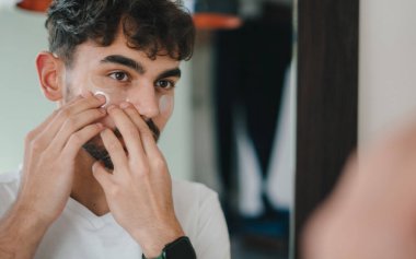 Genç bir adam sabah cilt bakımı sırasında aynanın önünde dikilirken özenle nemlendirici sürüyor.