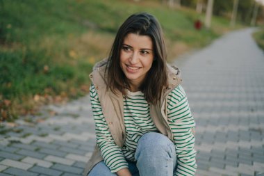 Genç bir kadın, doğa ile çevrili, rahat bir şekilde giyinmiş, bir patikada mutlu bir şekilde oturuyor.