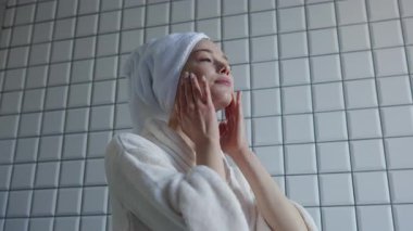 Bornoz ve havlu giyen bir kadın güzel tasarlanmış şık bir banyoda cilt bakımını yapıyor.