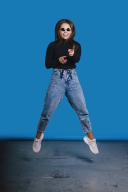 Şık güneş gözlüğü ve günlük kıyafetlerle havaya zıplayan bir kadının eğlenceli ve canlı bir görüntüsü.