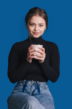 Huzurlu ve sakin genç bir kadın zarif bir şekilde elinde bir bardak tutarken canlı mavi bir fonda oturuyor.