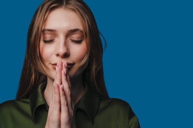 Genç bir kadın huzurlu bir şekilde, gözleri kapalı ve elleri mavi bir zemine yapışık bir şekilde derin derin düşünüyor.