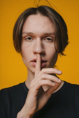 Genç bir adam, hem gizemi hem de entrikayı somutlaştıran parlak ve canlı sarı bir zemin karşısında sessizlik için işaret ediyor.